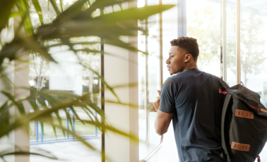 A young man wearing a backpack stands by a glass door, looking over his shoulder as he enters or exits a building. Sunlight filters through nearby greenery, creating a bright, modern indoor setting.