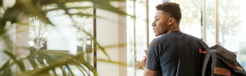 A young man wearing a backpack stands by a glass door, looking over his shoulder as he enters or exits a building. Sunlight filters through nearby greenery, creating a bright, modern indoor setting.